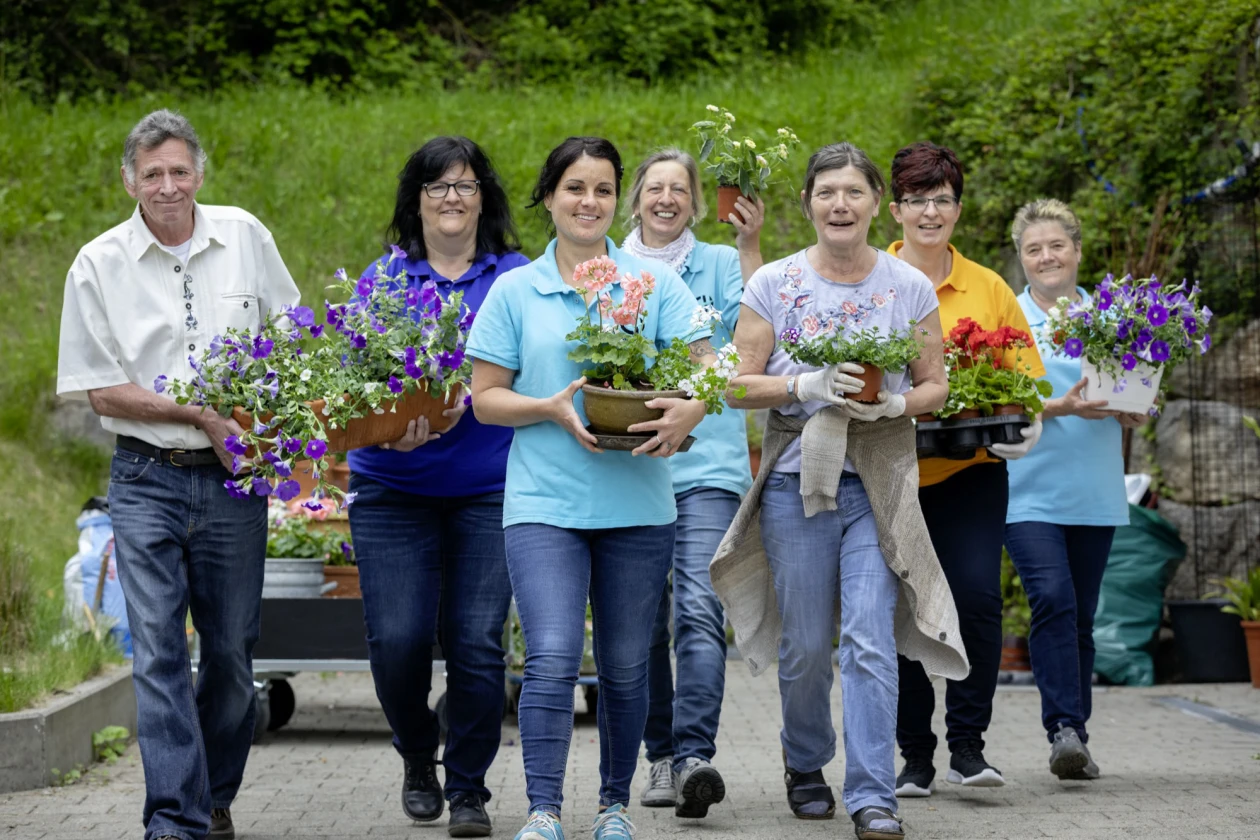 Sieben Personen gehen in einer Reihe, jeder tr&auml;gt einen Blumentopf und l&auml;chelt dabei. Im Hintergrund ist eine gr&uuml;ne Gartenumgebung zu sehen.