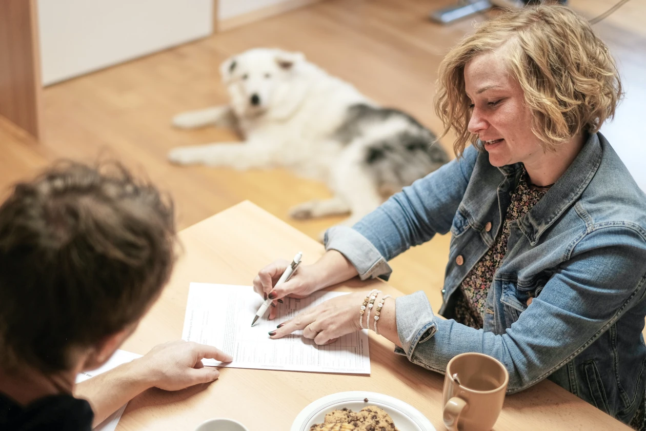 Zwei Personen sitzen an einem Tisch. Die Frau in Jeansjacke deutet mit einem Kugelschreiber auf ein Dokument. Ein wei&szlig;er Hund liegt im Hintergrund auf dem Boden. Auf dem Tisch stehen eine Tasse und eine Sch&uuml;ssel mit Keksen.