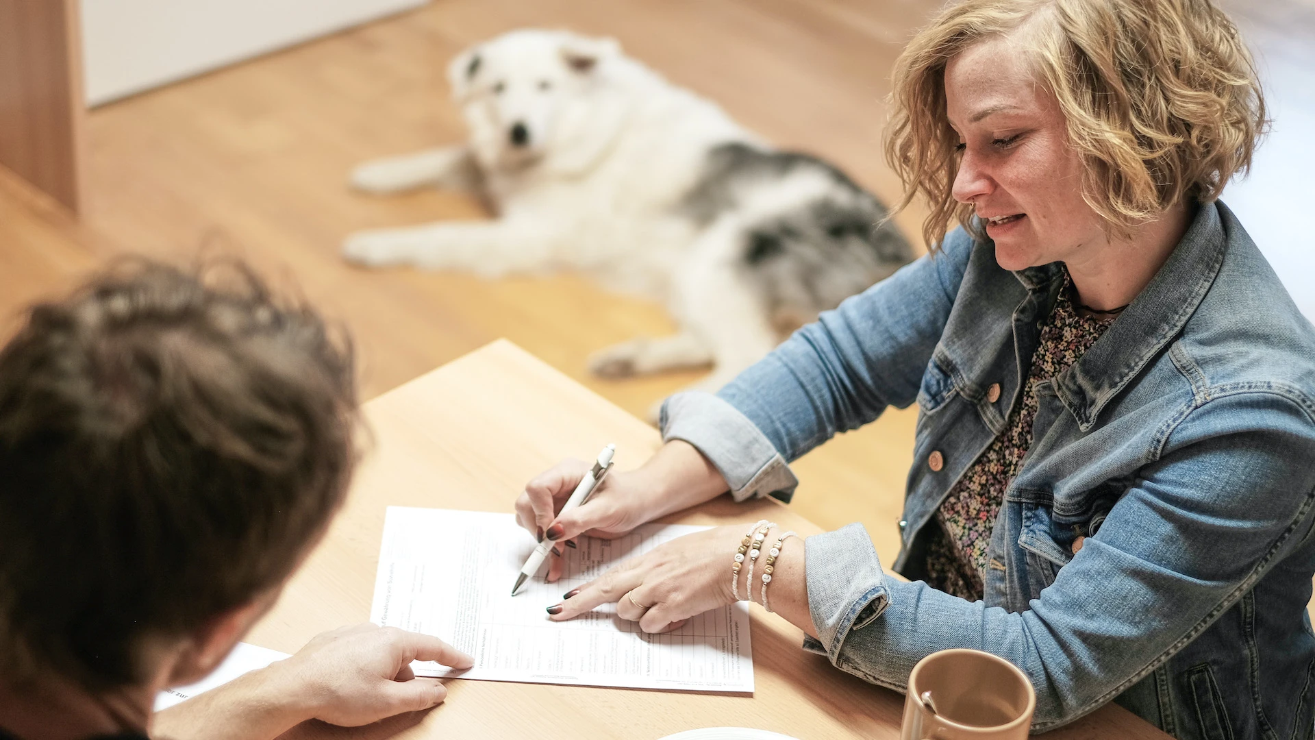 Zwei Personen sitzen an einem Tisch. Die Frau in Jeansjacke deutet mit einem Kugelschreiber auf ein Dokument. Ein wei&szlig;er Hund liegt im Hintergrund auf dem Boden. Auf dem Tisch stehen eine Tasse und eine Sch&uuml;ssel mit Keksen.