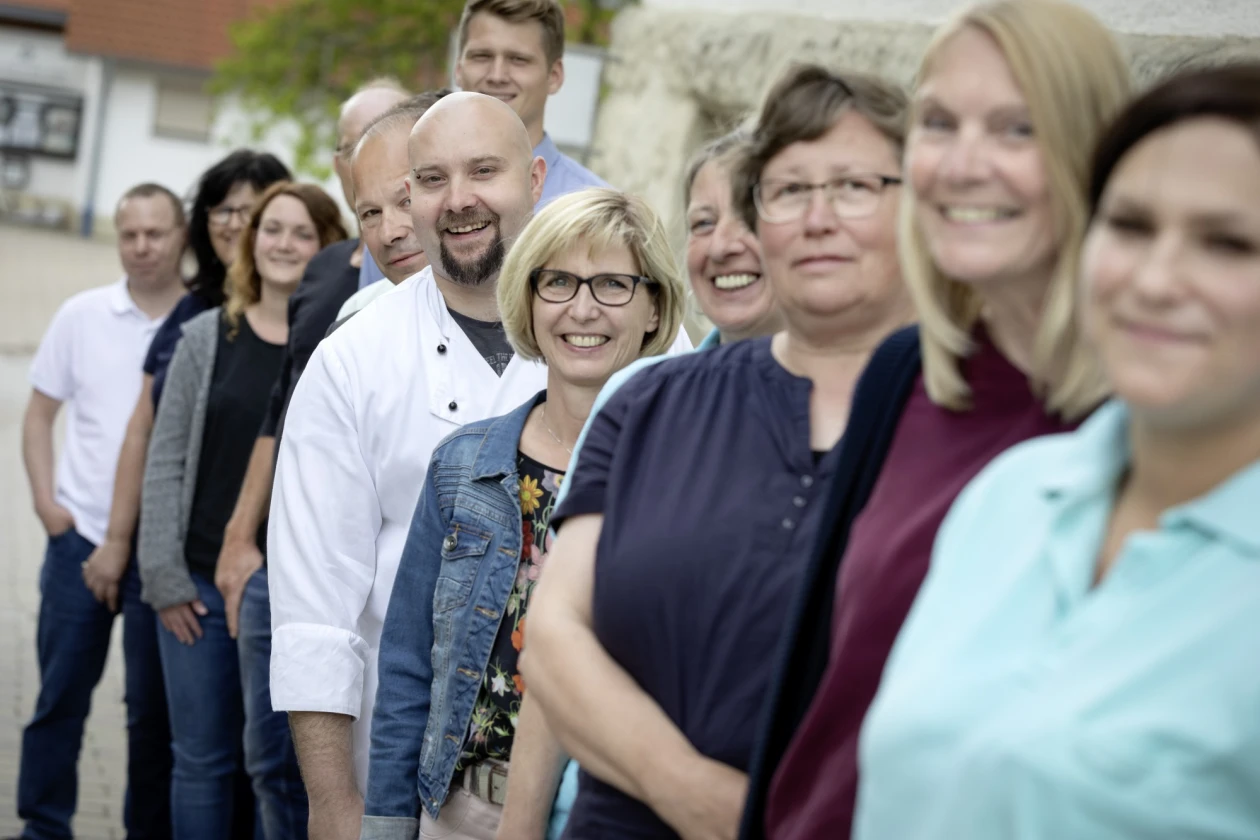 Eine Gruppe l&auml;chelnder Personen in unterschiedlicher Kleidung stehen hintereinander in einer Reihe an einer Steinmauer. Im Hintergrund ist ein wei&szlig;es Geb&auml;ude mit rotem Ziegeldach zu sehen.