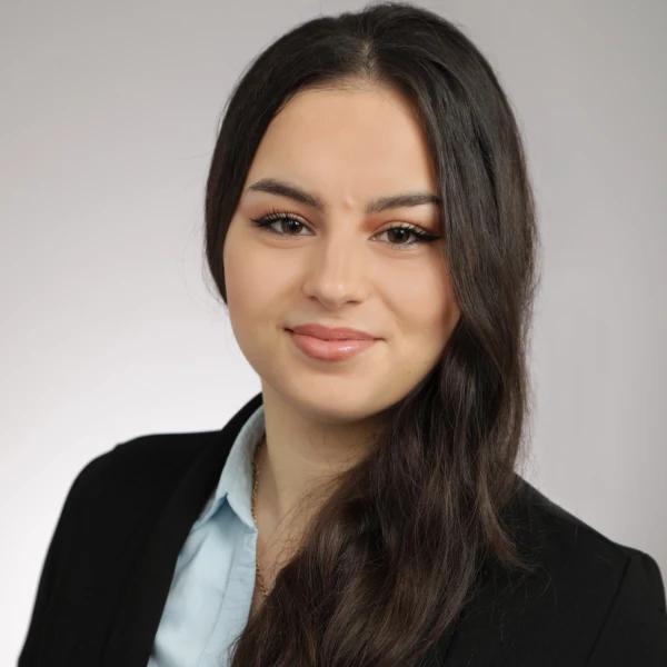 Junge Frau mit langen dunklen Haaren in einem schwarzen Blazer und hellblauer Bluse, die in die Kamera l&auml;chelt.