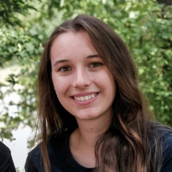 Eine junge, in die Kamera l&auml;chelnde Frau mit langen braunen Haaren. Sie tr&auml;gt ein dunkles Oberteil und befindet sich vor einem unscharfen, gr&uuml;nen Bl&auml;tterhintergrund.