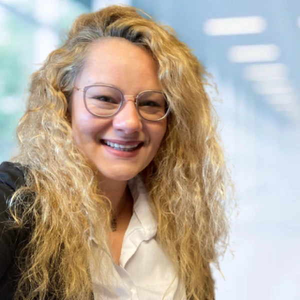 L&auml;chelnde Frau mit lockigem, blondem Haar und Brille in wei&szlig;er Bluse und schwarzem Blazer steht vor einem unscharfen, hellen Hintergrund.