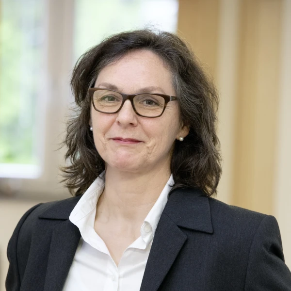 Eine l&auml;chelnde Frau mit schulterlangen braunen Haaren, mit Brille, gekleidet in einer wei&szlig;en Bluse mit schwarzem Blazer.