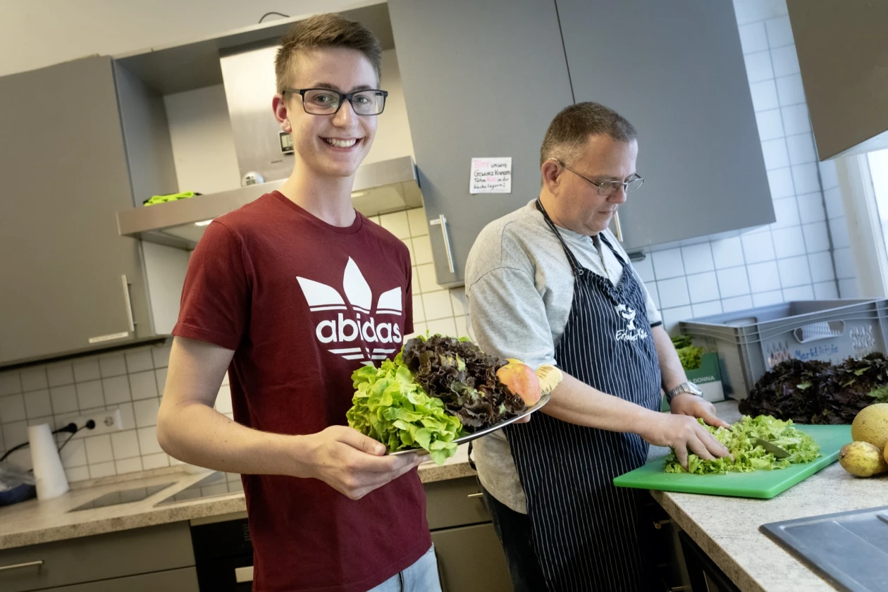 Zwei M&auml;nner in einer K&uuml;che, die frischen Salat zubereiten. Der j&uuml;ngere Mann in einem roten Adidas-T-Shirt l&auml;chelt in die Kamera und h&auml;lt einen gro&szlig;en Teller mit Salat in der Hand, w&auml;hrend der &auml;ltere Mann in einer gestreiften Sch&uuml;rze Salat auf einem gr&uuml;nen Schneidebrett schneidet.