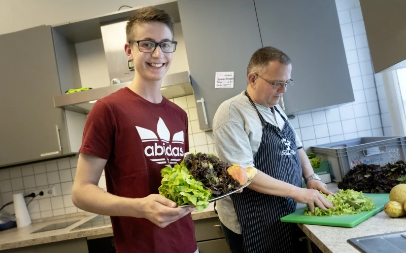 Zwei M&auml;nner in einer K&uuml;che, die frischen Salat zubereiten. Der j&uuml;ngere Mann in einem roten Adidas-T-Shirt l&auml;chelt in die Kamera und h&auml;lt einen gro&szlig;en Teller mit Salat in der Hand, w&auml;hrend der &auml;ltere Mann in einer gestreiften Sch&uuml;rze Salat auf einem gr&uuml;nen Schneidebrett schneidet.