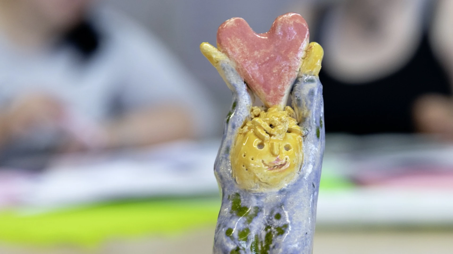 Handgefertigte Keramikfigur mit gelbem Gesicht und rosa Herz, in blau-gr&uuml;ne Glasur getaucht, die auf einem Holztisch vor verschwommenen Personen im Hintergrund steht.