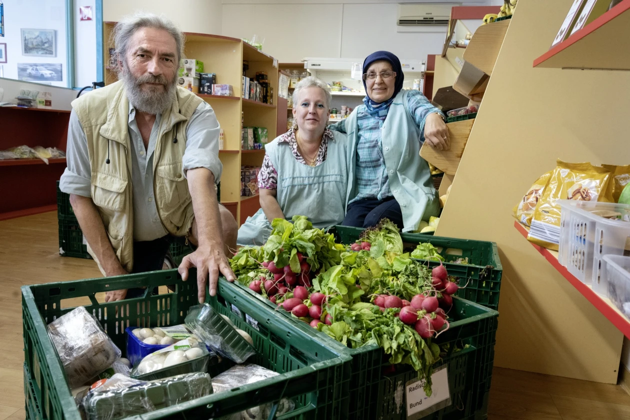 Drei Personen stehen hinter drei gr&uuml;nen Kisten mit Gem&uuml;se gef&uuml;llt. Links befindet sich ein b&auml;rtiger Mann mit Weste, in der Mitte steht eine Frau, rechts daneben eine weitere Frau mit Kopftuch. Im Hintergrund sind Regele mit Lebensmitteln zu sehen.