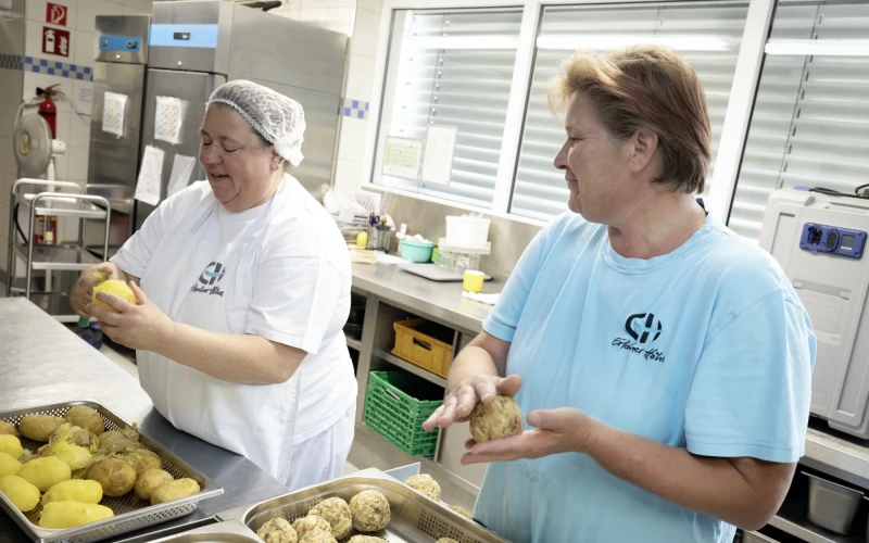 Zwei Frauen arbeiten in einer K&uuml;che. Eine Frau in wei&szlig;er Kleidung und Haube sch&auml;lt Kartoffeln, w&auml;hrend die andere in blauem T-Shirt Kn&ouml;del formt. Arbeitsumgebung ist sauber und professionell mit Edelstahlger&auml;ten und Ablageregalen.