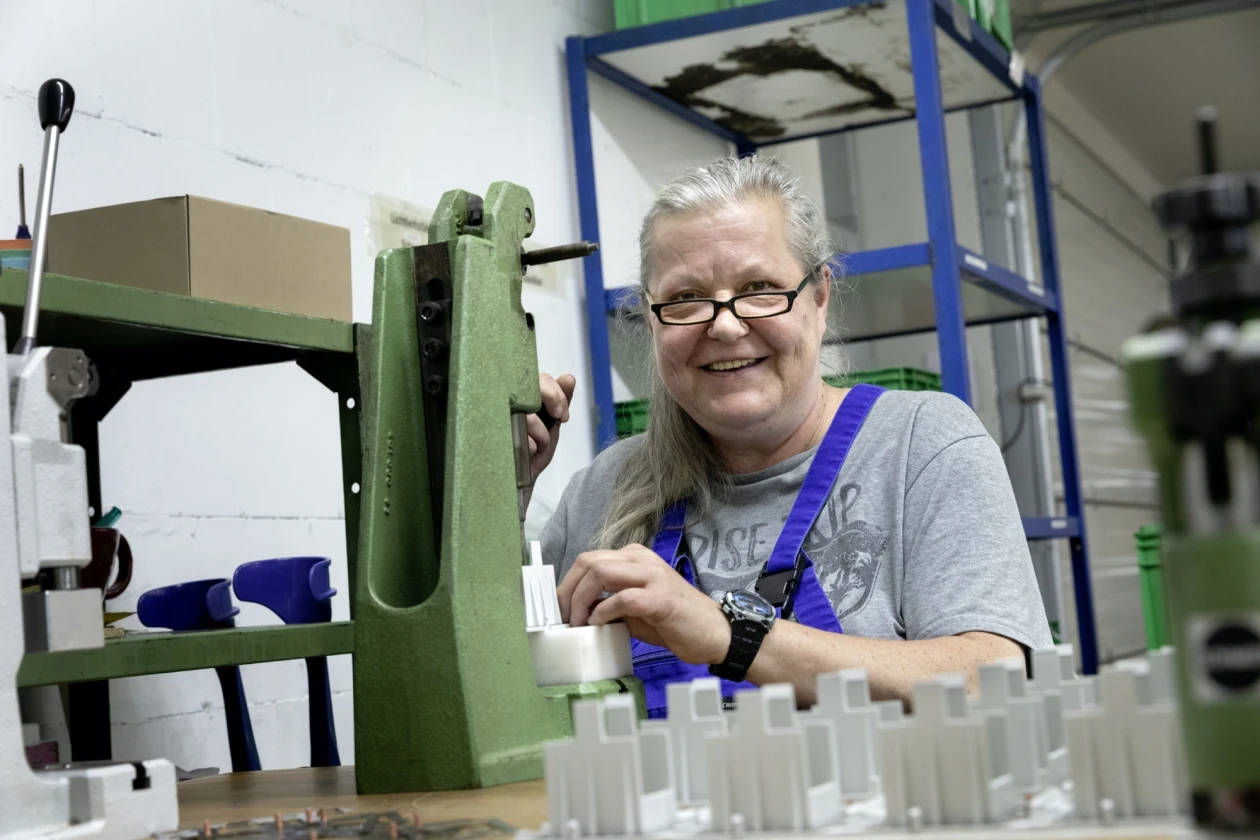 Eine &auml;ltere Frau mit Brille und grauem T-Shirt l&auml;chelt in die Kamera. Sie sitzt an einer gr&uuml;nen Maschine und legt Werkst&uuml;cke ein. Sie scheint zufrieden mit ihrer Arbeit zu sein. Die Umgebung zeigt verschiedene Maschinen und Regale im Hintergrund.