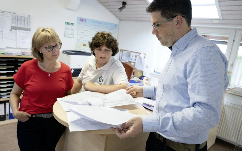 Drei Personen stehen an einem Tisch und betrachten gemeinsam einige Dokumente, die der Mann im hellblauen Hemd in den H&auml;nden h&auml;lt. Der Raum ist mit einem Wandkalender, Regalen voller Aktenordner, zwei Fenstern sowie weiterem B&uuml;romaterial und Unterlagen ausgestattet.