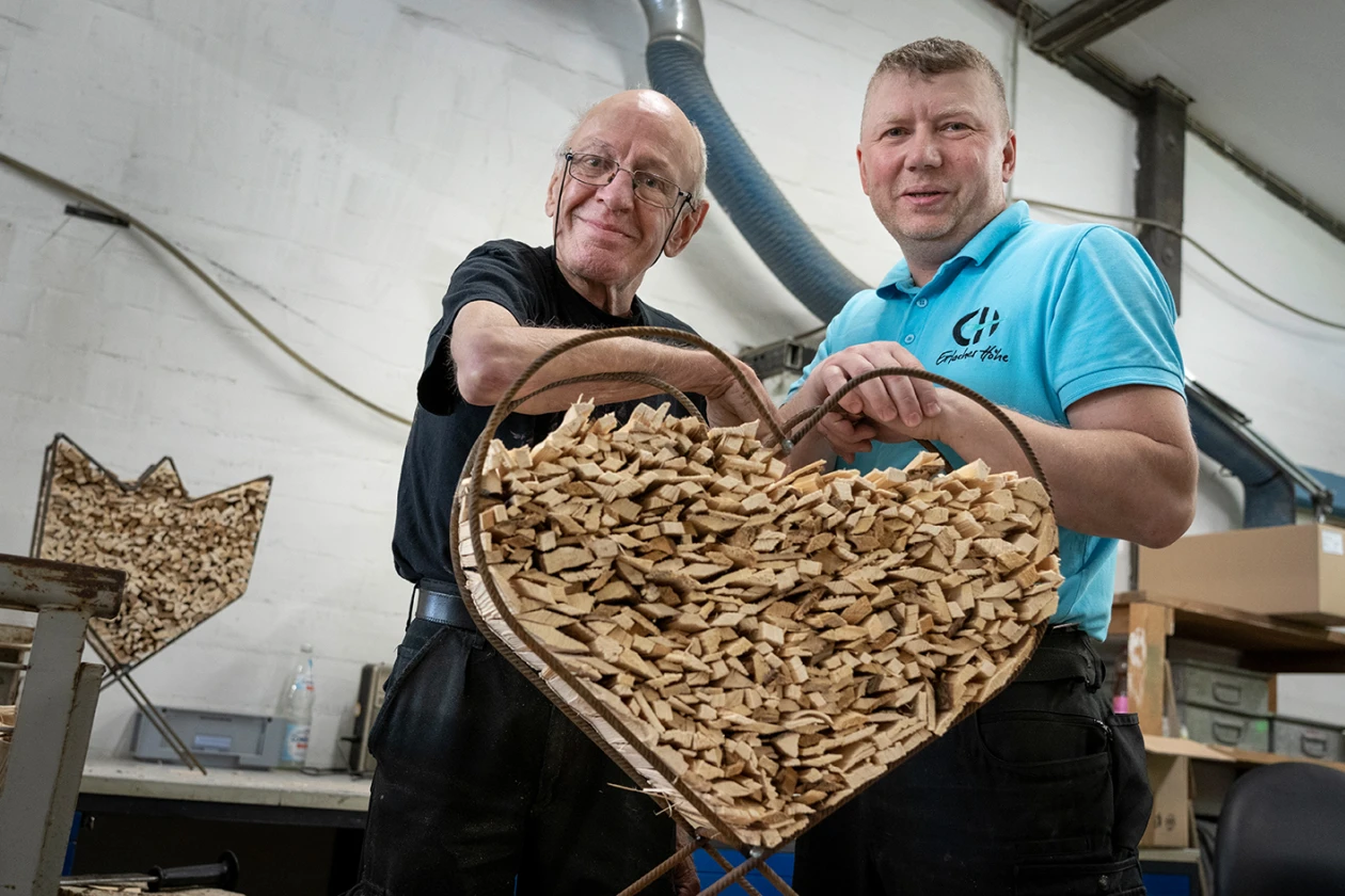 Zwei M&auml;nner halten gemeinsam ein gro&szlig;es, herzf&ouml;rmiges Metallgestell gef&uuml;llt mit Anz&uuml;ndholz. Der &auml;ltere Mann tr&auml;gt ein schwarzes T-Shirt, der j&uuml;ngere ein hellblaues T-Shirt mit Logo.